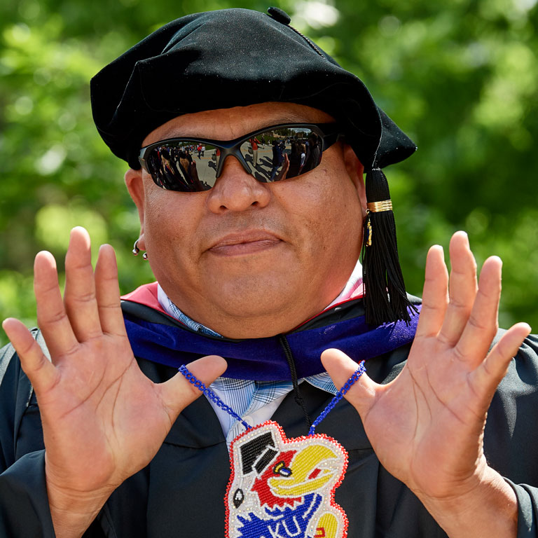 A law student in graduation regalia holds up a Jayhawk necklace