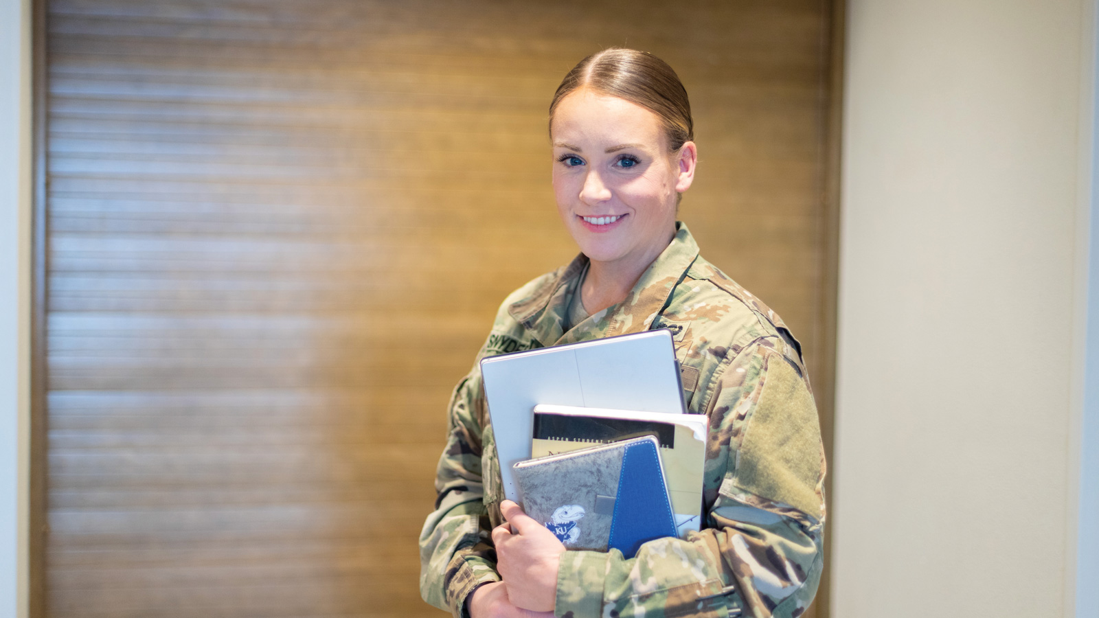 A female student in military fatigues holds textbooks and notebooks.